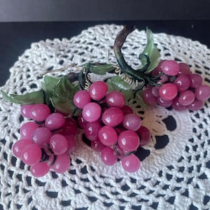 Puede incluir: Un conjunto decorativo de tres racimos de uvas rosas hechos de piedras lisas y redondeadas. Cada racimo presenta uvas esféricas de color rosa translúcido unidas a tallos de alambre verde oscuro con hojas de piedra verde tallada.
