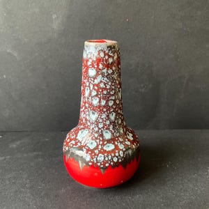 Op de afbeelding: Een kleine keramische vaas met een bolvormige basis en een taps toelopende hals. Het oppervlak heeft een felrood glazuur aan de onderkant, dat naar boven toe overgaat in een textuurrijk, gespikkeld patroon van wit en grijs op een dieprode achtergrond.