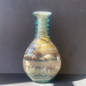 Könnte beinhalten: Eine klare Glasflasche mit engem Hals und spiralförmigem Glasdesign. Das Innere ist mit geschichtetem Sand gefüllt, der eine Wüstenlandschaft mit drei Kamelen am Boden und Bergspitzen im Hintergrund darstellt.