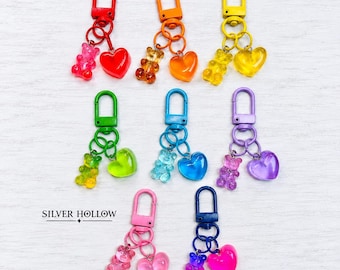 Pode incluir: Oito porta-chaves coloridos com berloques de ursinhos de goma translúcidos e berloques em forma de coração presos a mosquetões de metal correspondentes. O texto SILVER HOLLOW está impresso a preto à esquerda. Cada conjunto tem uma cor.