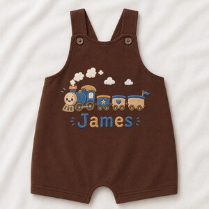 May include: A dark brown overall romper featuring an embroidered blue and tan train with white smoke clouds. The name James is embroidered in blue below the train. The garment has two shoulder straps secured with brown buttons at the front.