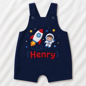 May include: A navy blue romper featuring an embroidered space theme with a white and red rocket, a smiling astronaut, and yellow and blue stars. The name Henry is embroidered in red letters below the space design. The romper has two shoulder buttons.