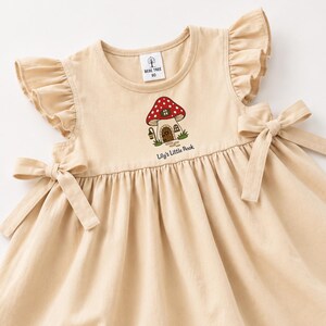 Pode incluir: Um vestido bege com mangas de folhos e laços laterais na cintura. O peito apresenta um gráfico bordado de uma casa cogumelo vermelha e branca com uma lanterna e o texto Lily's Little Book. A etiqueta no pescoço diz Bebe Tree 80.