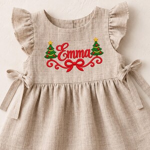 Puede incluir: Un vestido beige sin mangas con hombros con volantes y lazos laterales. El pecho presenta un diseño bordado con el nombre Emma en letra roja, flanqueado por dos árboles de Navidad verdes con estrellas amarillas y un lazo rojo debajo.