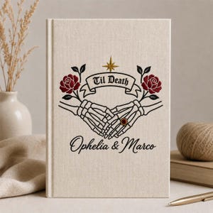 May include: A beige fabric-covered book featuring an embroidered design of two skeleton hands holding each other, with red roses, a gold star, and a banner reading Til Death. Below the hands, the names Ophelia & Marco are embroidered in black.