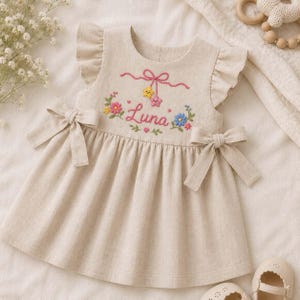 May include: A beige sleeveless dress with ruffled shoulders and side bows. The bodice features embroidered flowers, a pink bow, a yellow star, a pink star, and the name Luna in pink script. The dress has a round neckline and a gathered skirt.