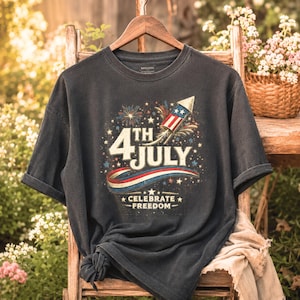 May include: Dark gray t-shirt with a distressed graphic celebrating the 4th of July. The design features a rocket, fireworks, and the text "4th July" and "Celebrate Freedom" in red, white, and blue.