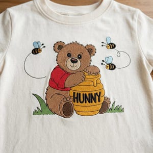 May include: An off-white t-shirt featuring an embroidered design of a brown bear wearing a red shirt, hugging a yellow honey pot labeled HUNNY. The design includes two small flying bees with blue wings and green grass at the base of the bear.