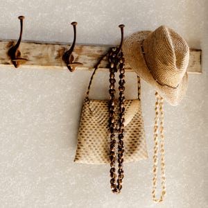 Può includere: Una borsa a mano in paglia intrecciata marrone chiaro con motivo geometrico, una lunga tracolla di perline e un cappello fedora in paglia abbinato con una collana di conchiglie chiara appesa alla tesa, tutto appeso a un appendiabiti da parete in legno.