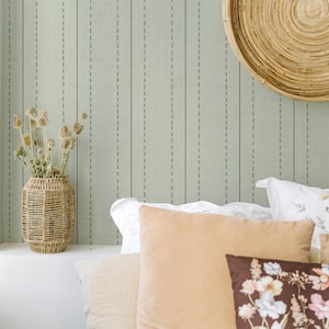 Op de afbeelding: Een saliegroen behang met verticale stippellijnen, een gevlochten rotan vaas met droogbloemen, een ronde wandhanger van riet en diverse kussens in wit, beige en bruin, waarbij het voorste kussen is voorzien van een bloemenprint.