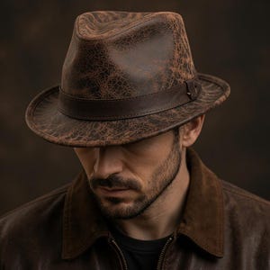 Puede incluir: Un sombrero fedora marrón con una textura agrietada y desgastada, con una banda de cuero marrón oscuro alrededor de la copa. El sombrero presenta una copa pellizcada y un ala estructurada, mostrando un aspecto envejecido en tonos marrones.