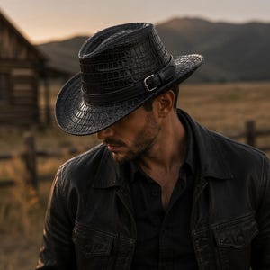 Könnte beinhalten: Ein schwarzer Fedora-Hut mit einer strukturierten Krokodilleder-Optik und einem passenden schwarzen Lederband mit einer silberfarbenen Metallschnalle. Der Hut hat eine strukturierte Krone und eine breite Krempe, was ihm ein markantes Aussehen verleiht.
