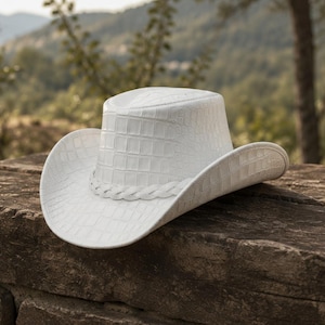 May include: A white cowboy hat featuring a textured crocodile skin pattern across the crown and brim. The hat includes a braided white band encircling the base of the crown, with a curved brim and a structured, indented top design.