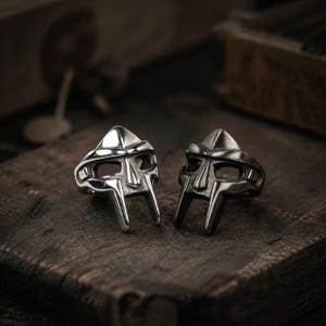 Puede incluir: Dos anillos de metal con forma de máscaras de gladiador. Un anillo tiene un acabado plateado brillante y pulido, mientras que el otro presenta un acabado metálico más oscuro, cepillado u oxidado. Ambos anillos muestran detalles faciales.