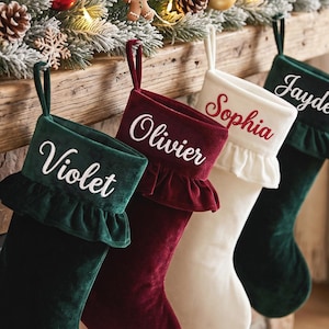 May include: Four velvet Christmas stockings with ruffled cuffs and hanging loops. The stockings are dark green, deep red, and cream. Each cuff features embroidered names: Violet in white, Olivier in white, Sophia in red, and Jayde in white.