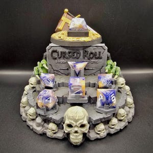 Puede incluir: Un soporte de exhibición gris de varios niveles con una calavera en la base y el texto Cursed Roll en el panel posterior. El soporte sostiene un juego de siete dados poliédricos con un patrón jaspeado blanco, azul y naranja.