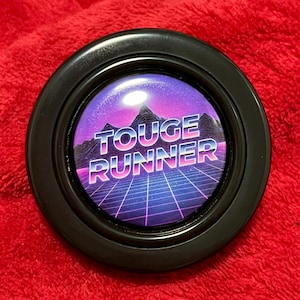 Könnte beinhalten: Ein schwarzer, kreisförmiger Knopf mit dem Text "TOUGE RUNNER" in Weiß und Rosa, vor einer Retro-Landschaft aus Bergen und einem Raster. Der Knopf hat eine glänzende Oberfläche und liegt auf einer roten Oberfläche.
