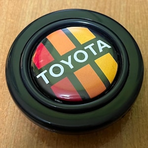 May include: A black Toyota emblem with a colorful design. The emblem features the Toyota logo in white, set against a backdrop of red, orange, and yellow rectangles. The emblem is circular and has a glossy finish.