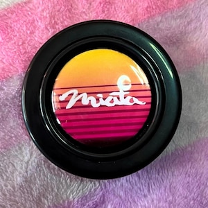Puede incluir: Un estuche compacto redondo y negro con un diseño de puesta de sol en tonos naranja, amarillo y rosa. La palabra "Miata" está escrita en blanco en el centro del diseño. El estuche compacto está sobre una superficie rosa y morada.