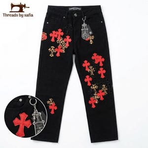 May include: Black denim jeans featuring multiple red cross-shaped patches and leopard print cross-shaped patches scattered across the front. The jeans include a silver-toned metal ring attached to the front belt loop with a branded tag.