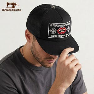 May include: A black trucker hat featuring a rectangular patch on the front. The patch displays the text CHROME HEARTS and HOLLYWOOD, USA, with a graphic of red lips and a silver cross emblem. The hat has a mesh back and a silver button on top.