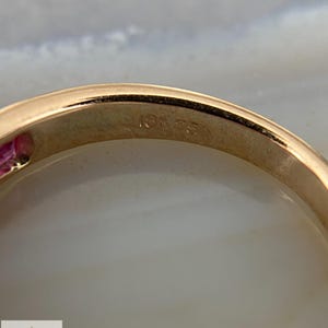 May include: A gold ring featuring a smooth, polished band with the hallmark 18K 750 engraved on the interior surface. A small portion of a pink gemstone is visible on the left side of the band, which has a rounded profile and a metallic finish.