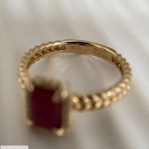 May include: A gold ring featuring a rectangular red gemstone and a textured, braided-style band. The interior of the band is engraved with the text 14K IDJ, RD0.077, and RUB1.210, indicating the gold purity and the weight of the stones in carats.