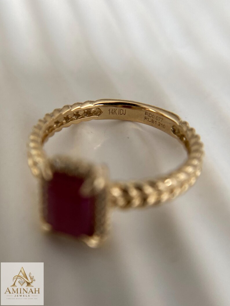 May include: A gold ring featuring a rectangular red gemstone and a textured, braided-style band. The interior of the band is engraved with the text 14K IDJ, RD0.077, and RUB1.210, indicating the gold purity and the weight of the stones in carats.