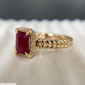 May include: A gold ring featuring a rectangular, deep red ruby center stone surrounded by a halo of small, clear accent stones. The ring band has a textured, braided design. The logo for Aminah Jewels is visible in the bottom left corner.