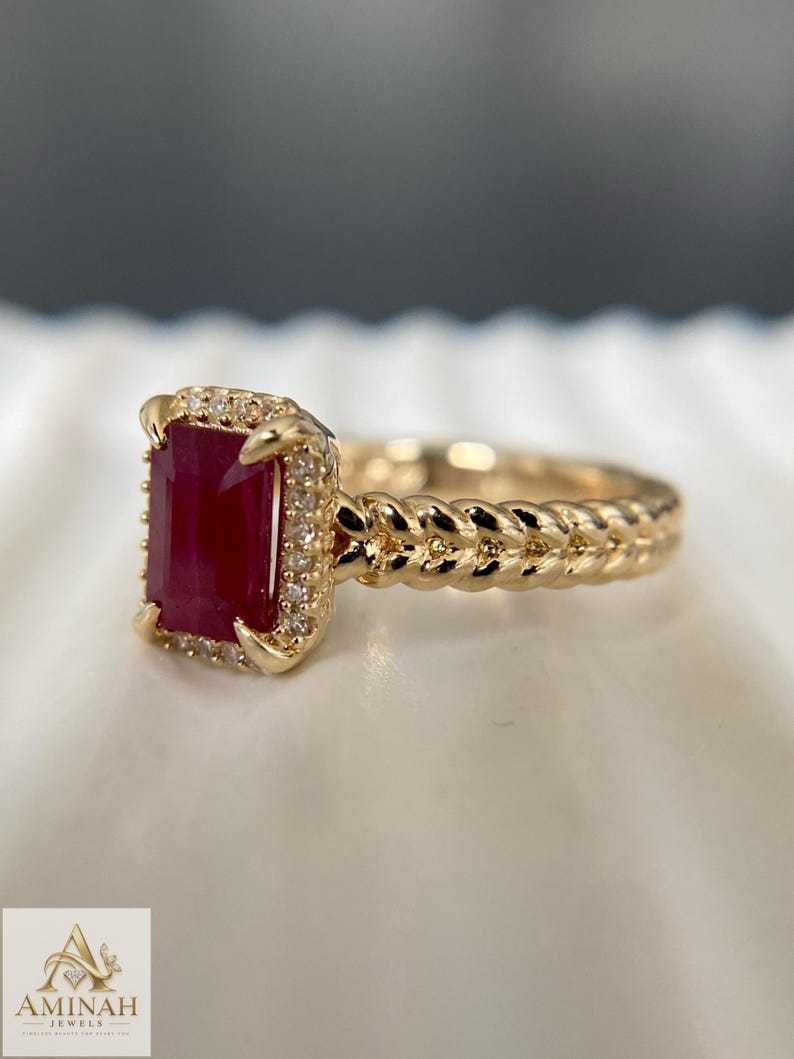 May include: A gold ring featuring a rectangular, deep red ruby center stone surrounded by a halo of small, clear accent stones. The ring band has a textured, braided design. The logo for Aminah Jewels is visible in the bottom left corner.