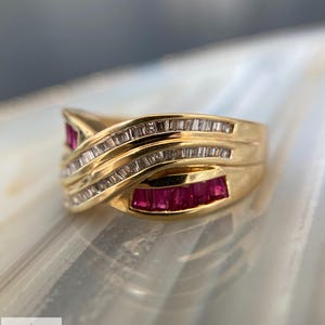 K&ouml;nnte beinhalten: Ein Goldring mit &uuml;berkreuztem Design, besetzt mit im Kanal gefassten Baguette-Rubinen und Reihen von im Kanal gefassten Baguette-Diamanten. Der Ring hat eine polierte Oberfl&auml;che und eine geschwungene Struktur, die die roten und klaren Steine betont.