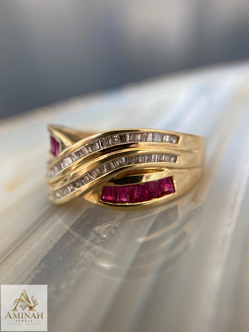 K&ouml;nnte beinhalten: Ein Goldring mit &uuml;berkreuztem Design, besetzt mit im Kanal gefassten Baguette-Rubinen und Reihen von im Kanal gefassten Baguette-Diamanten. Der Ring hat eine polierte Oberfl&auml;che und eine geschwungene Struktur, die die roten und klaren Steine betont.