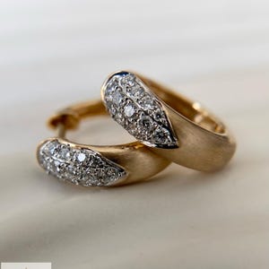 Könnte beinhalten: Ein Paar goldene Creolen mit einer gebürsteten, matten Oberfläche am Ring und einem tropfenförmigen Abschnitt, der mit kleinen, klaren, funkelnden Steinen besetzt ist. Die Ohrringe sind zusammen positioniert und zeigen den Kontrast zwischen dem texturierten Gold und den hellen, reflektierenden Steinen. Das Logo von Aminah Jewels ist in der unteren Ecke zu sehen.