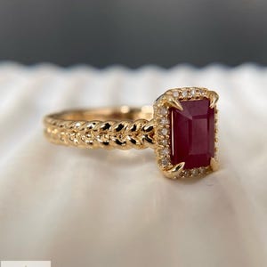 May include: A gold ring featuring a rectangular deep red gemstone set in a halo of small white stones. The band has a braided, rope-like texture. The logo for Aminah Jewels is visible in the corner, featuring a stylized letter A and text.