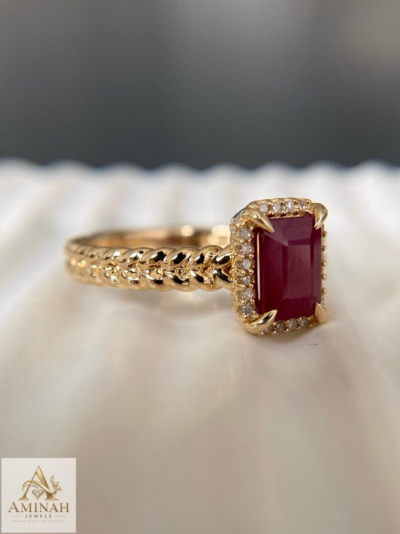 May include: A gold ring featuring a rectangular deep red gemstone set in a halo of small white stones. The band has a braided, rope-like texture. The logo for Aminah Jewels is visible in the corner, featuring a stylized letter A and text.