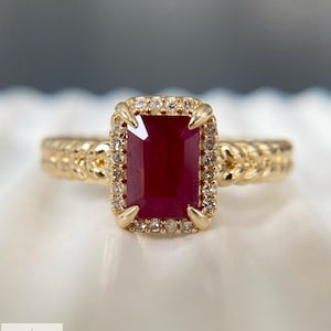 May include: A gold ring featuring a rectangular emerald-cut deep red ruby center stone surrounded by a halo of small round white diamonds. The ring band has a textured braided design. The logo Aminah Jewels is visible in the bottom corner.