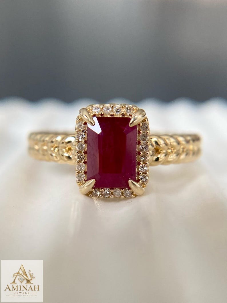 May include: A gold ring featuring a rectangular emerald-cut deep red ruby center stone surrounded by a halo of small round white diamonds. The ring band has a textured braided design. The logo Aminah Jewels is visible in the bottom corner.
