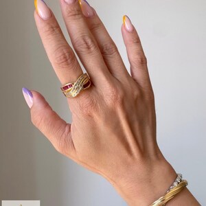 K&ouml;nnte beinhalten: Ein Goldring mit &uuml;berkreuztem Design, besetzt mit im Kanal gefassten, baguettegeschliffenen roten Rubinen und runden wei&szlig;en Diamanten. Ein gedrehter Goldreif und ein silberfarbenes Armband mit runden wei&szlig;en Steinen werden am Handgelenk getragen. Das Logo AMINAH JEWELS ist sichtbar.