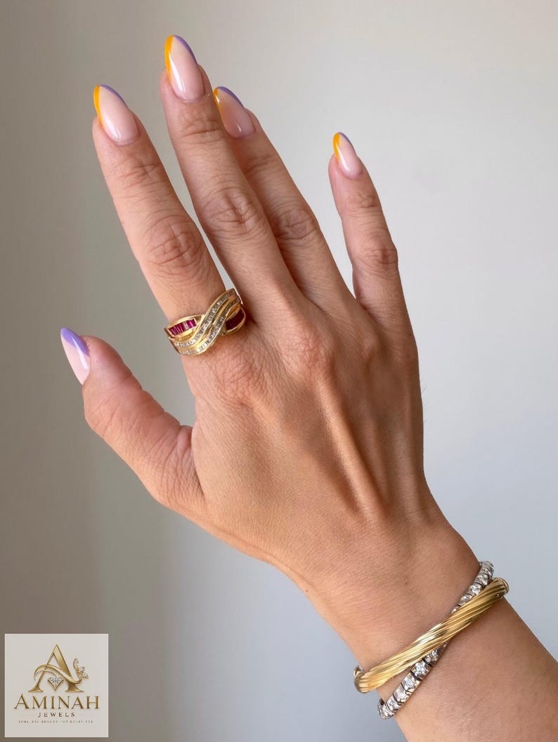 K&ouml;nnte beinhalten: Ein Goldring mit &uuml;berkreuztem Design, besetzt mit im Kanal gefassten, baguettegeschliffenen roten Rubinen und runden wei&szlig;en Diamanten. Ein gedrehter Goldreif und ein silberfarbenes Armband mit runden wei&szlig;en Steinen werden am Handgelenk getragen. Das Logo AMINAH JEWELS ist sichtbar.