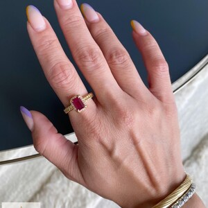 May include: A gold ring featuring a rectangular red gemstone surrounded by a halo of small white stones on a textured band, paired with a gold twisted bangle and a silver-toned tennis bracelet with clear stones, featuring the Aminah Jewels logo.