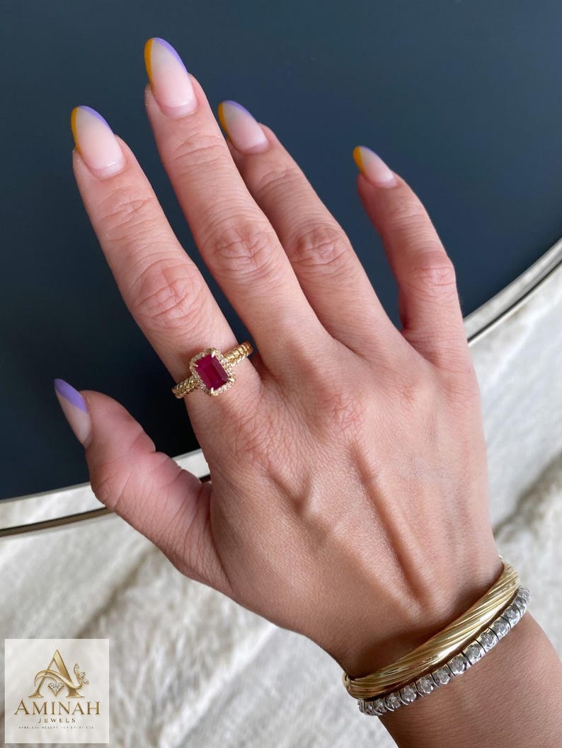 May include: A gold ring featuring a rectangular red gemstone surrounded by a halo of small white stones on a textured band, paired with a gold twisted bangle and a silver-toned tennis bracelet with clear stones, featuring the Aminah Jewels logo.