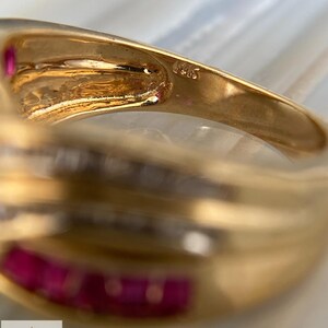 K&ouml;nnte beinhalten: Ein Goldring mit einer in Kanalfassung gesetzten Reihe aus rechteckigen roten Edelsteinen und einer Reihe klarer Steine. Die Innenseite der Ringschiene ist mit dem Text 14K gepr&auml;gt. Der Ring hat eine polierte Oberfl&auml;che und ein geschwungenes Design.