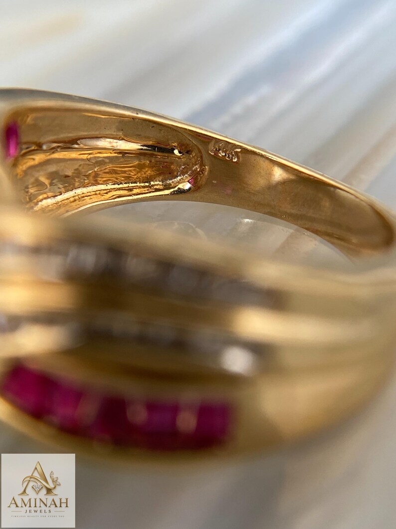 K&ouml;nnte beinhalten: Ein Goldring mit einer in Kanalfassung gesetzten Reihe aus rechteckigen roten Edelsteinen und einer Reihe klarer Steine. Die Innenseite der Ringschiene ist mit dem Text 14K gepr&auml;gt. Der Ring hat eine polierte Oberfl&auml;che und ein geschwungenes Design.