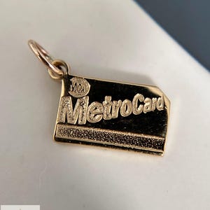 Könnte beinhalten: Ein goldfarbener Metallanhänger in Form einer rechteckigen MetroCard. Der Anhänger zeigt den geprägten Schriftzug MetroCard und das MTA-Logo in der oberen linken Ecke. Er hat einen texturierten unteren Bereich und einen kleinen runden Ring oben.