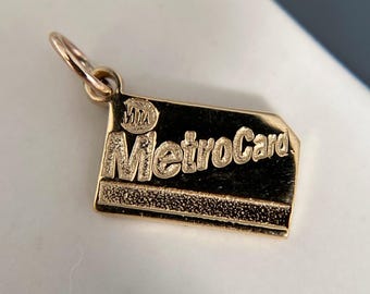 May include: A gold-toned metal charm shaped like a rectangular MetroCard. The charm features the embossed text MetroCard and the MTA logo in the upper left corner. It has a textured lower section and a small circular jump ring attached at the top.
