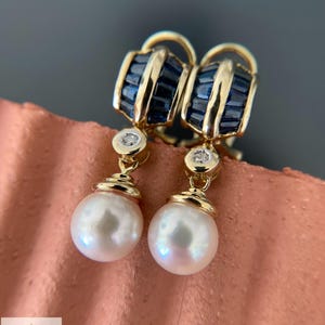 Könnte beinhalten: Ein Paar goldene Ohrringe mit einem oberen Abschnitt aus kanalgefassten Saphiren im Baguetteschliff, einem kleinen runden Diamantakzent und einer hängenden weißen Perle. Das Logo von Aminah Jewels ist unten links im Bild zu sehen.
