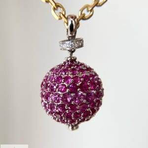 Könnte beinhalten: Ein kugelförmiger Anhänger, der mit runden, leuchtend pinken Edelsteinen besetzt ist und an einer Goldkette hängt. Der Anhänger hat eine silberfarbene Öse, die mit kleinen, klaren Pavé-Steinen verziert ist. Das Logo von Aminah Jewels ist sichtbar.
