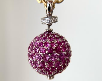 May include: A spherical pendant covered in round, vibrant pink gemstones, suspended from a gold chain. The pendant features a silver-toned bail accented with small, clear pavé-set stones. The logo for Aminah Jewels is visible in the bottom corner.