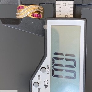 K&ouml;nnte beinhalten: Ein goldener Crossover-Ring mit im Kanal gefassten Baguette-Rubinen und Reihen kleiner runder Diamanten. Der Ring liegt in einer schwarzen digitalen Messschieber-Lehre, die auf dem Display 10,0 mm anzeigt, was die Breite des Rings angibt.