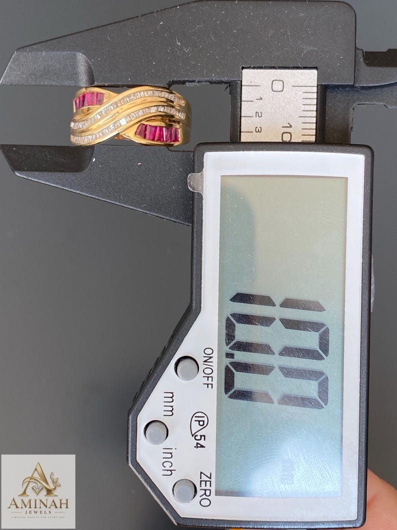 K&ouml;nnte beinhalten: Ein goldener Crossover-Ring mit im Kanal gefassten Baguette-Rubinen und Reihen kleiner runder Diamanten. Der Ring liegt in einer schwarzen digitalen Messschieber-Lehre, die auf dem Display 10,0 mm anzeigt, was die Breite des Rings angibt.